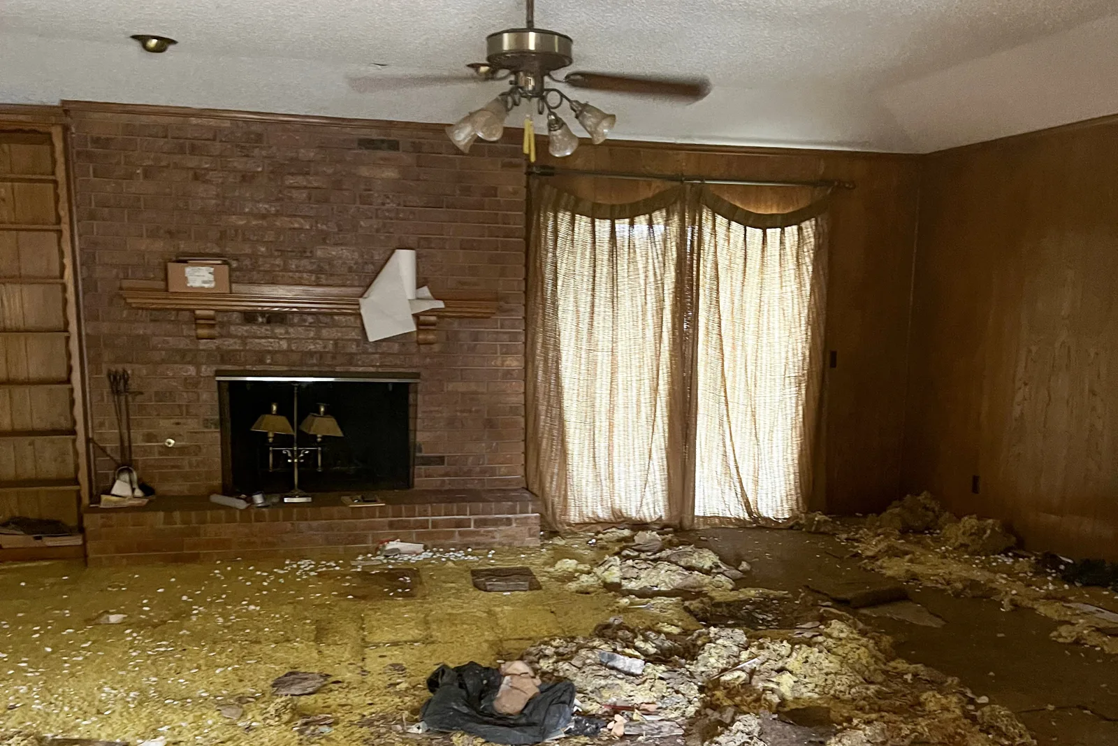 Plano living room before renovation, with the original red brick fireplace, wood wall panelling, and a bare subfloor covered in torn-out insulation and debris.