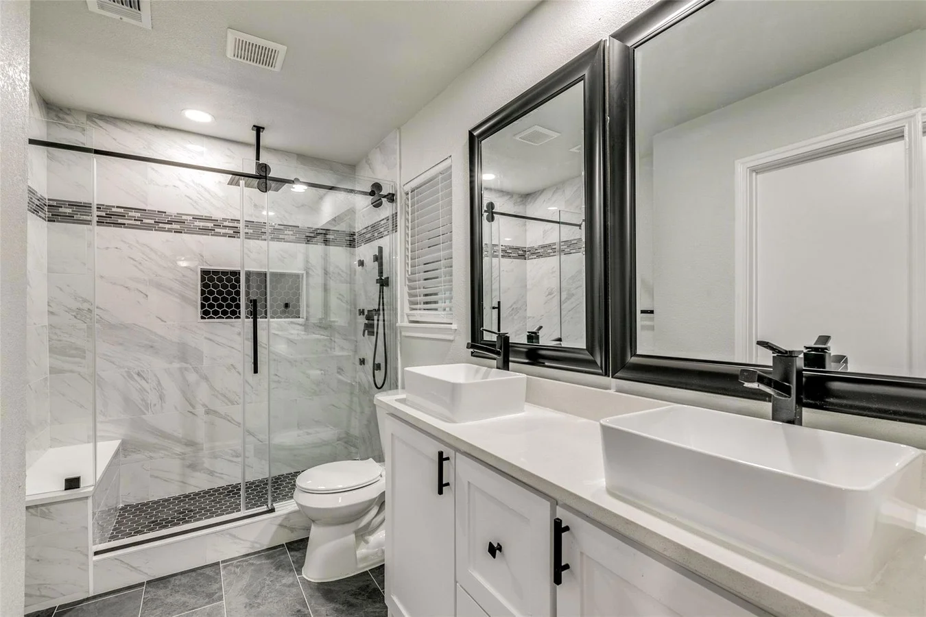Primary bathroom with a frameless glass shower, marble-look tile, double vessel vanity, black fixtures, and broad mirrors in a completed Plano, Texas whole-home remodel.