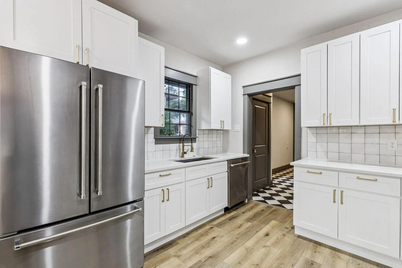 White kitchen with shaker cabinetry, stainless steel refrigerator, brass hardware, subway tile, and a checkerboard utility room beyond in Bishop Arts, Dallas, Texas.