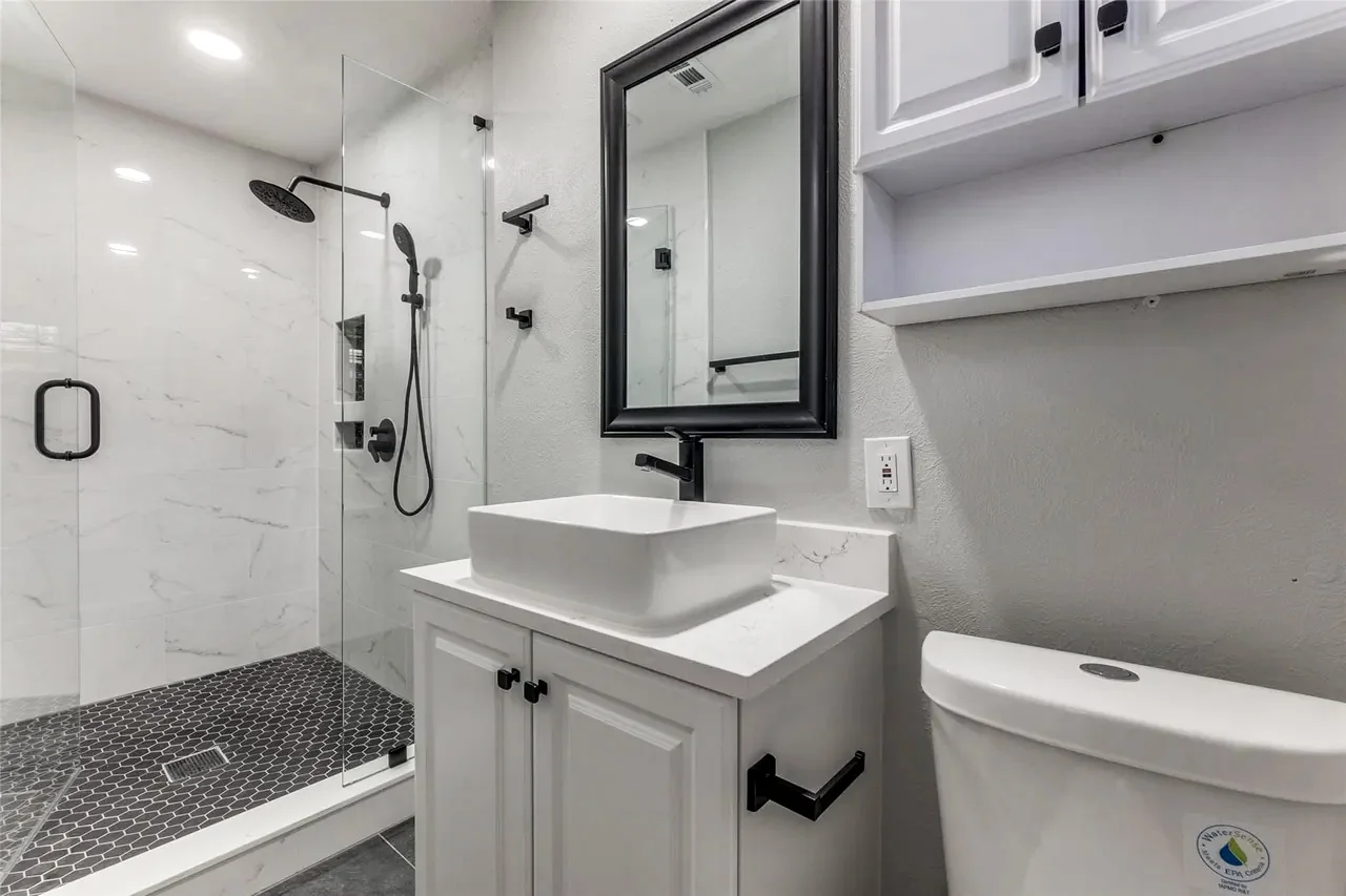 Primary bathroom with compact dark vanity, black fixtures, glass shower, and wood-look tile floor in Dallas.