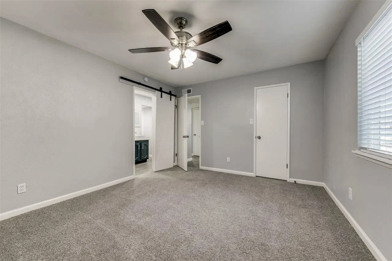 Primary bedroom with gray carpet, light walls, white trim, ceiling fan, and a sliding barn door to the bathroom in Dallas, Texas.