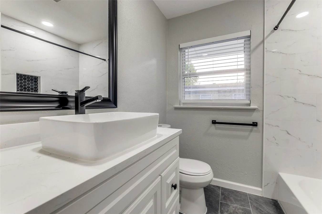 Guest bathroom with a white vessel sink, black fixtures, gray vanity, marble-look tub surround, and recessed lighting in Dallas, Texas.