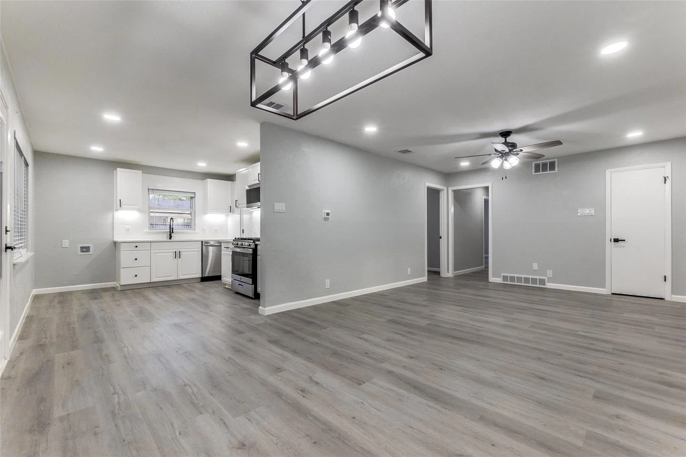 Open kitchen and living area with white cabinetry, gray wood-look flooring, recessed lighting, and a linear chandelier in Dallas, Texas.