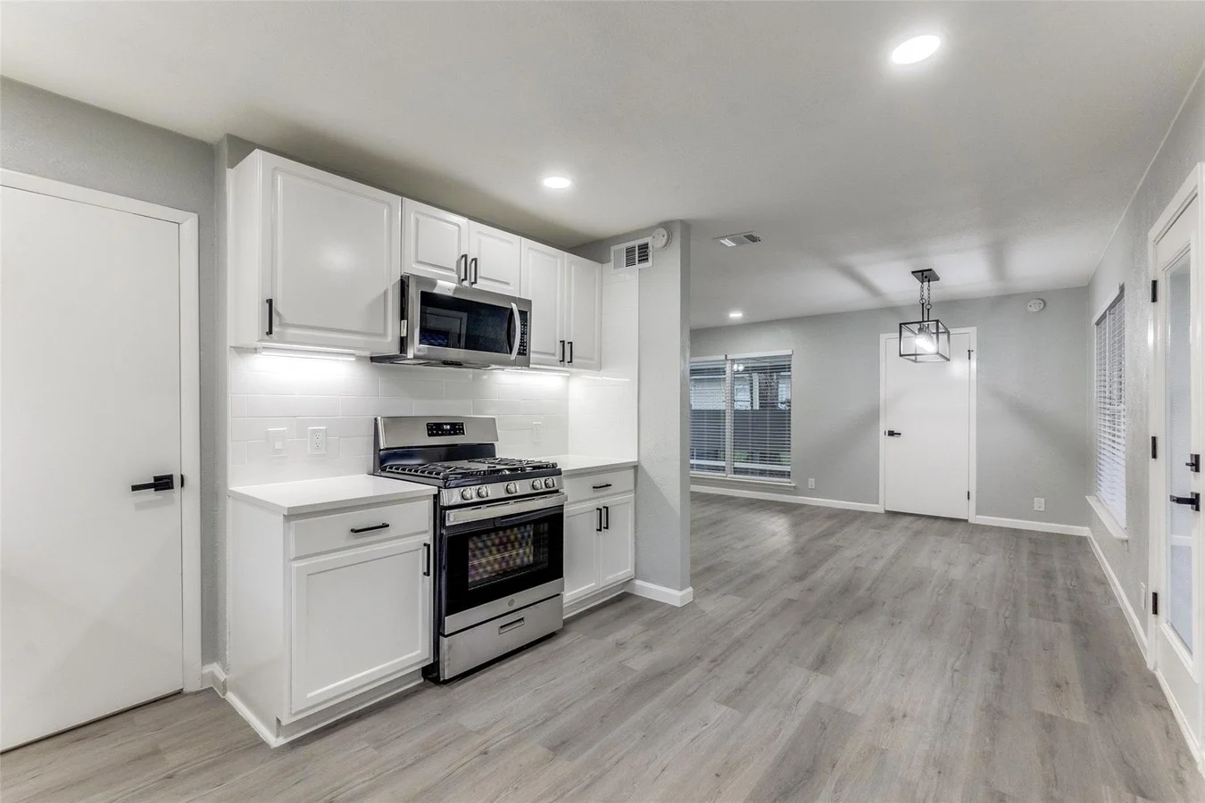 White kitchen opening to the dining and living areas with stainless appliances, recessed lighting, and gray wood-look flooring in Dallas, Texas.