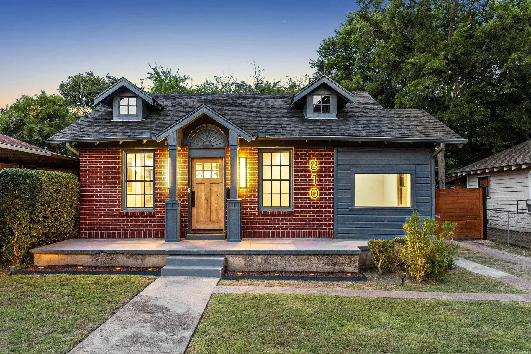 Painted Dallas cottage exterior with dark siding, warm wood door, lit windows, and landscaped front walk.