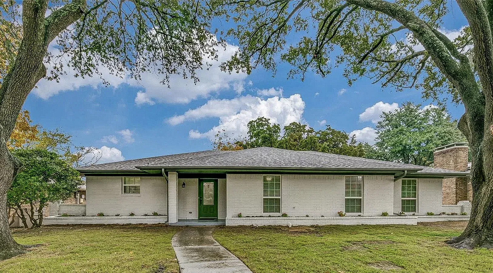 Plano ranch home with light exterior paint, dark shutters, green front door, and mature trees.