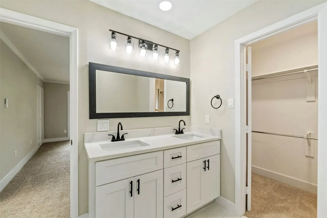 Primary bathroom double vanity with white cabinetry, black fixtures, broad mirror, recessed lighting, and walk-in closets in Fort Worth, Texas.