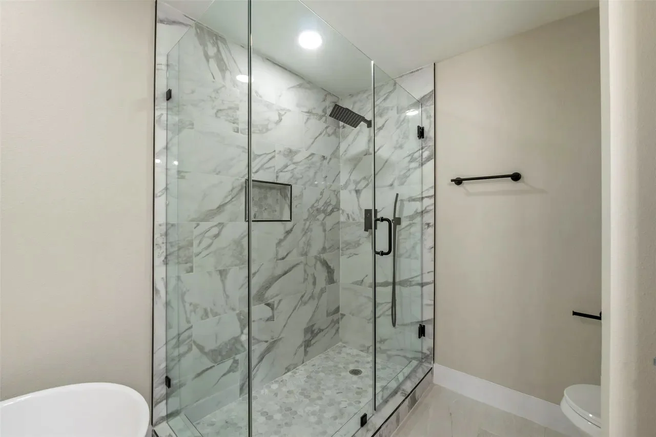 Primary bathroom shower with marble-look tile, frameless glass, black fixtures, built-in niche, and neutral flooring in Fort Worth, Texas.
