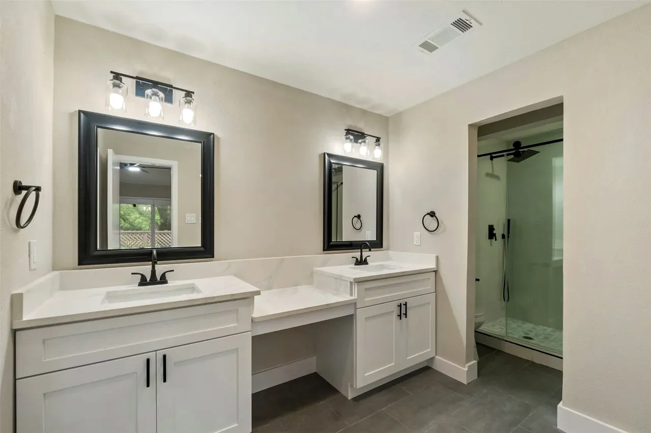 Second primary suite bathroom with a double vanity, makeup desk, black mirrors, dark flooring, and adjacent glass shower in Fort Worth, Texas.