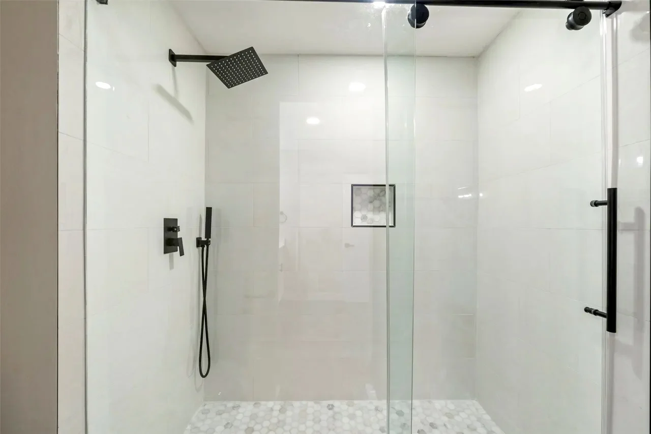 Primary suite shower with white tile, frameless glass, black rain shower fixtures, and pebble-look shower floor in Fort Worth, Texas.