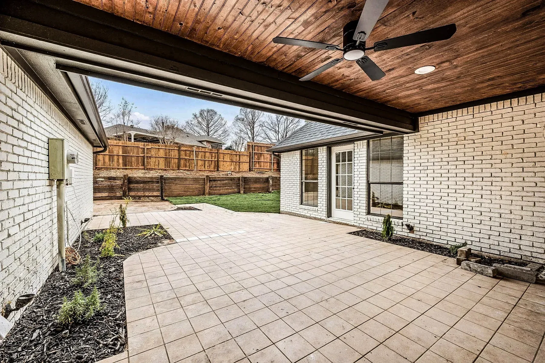 Covered Grapevine patio with stained wood ceiling, paver courtyard, white brick walls, and landscaped beds.