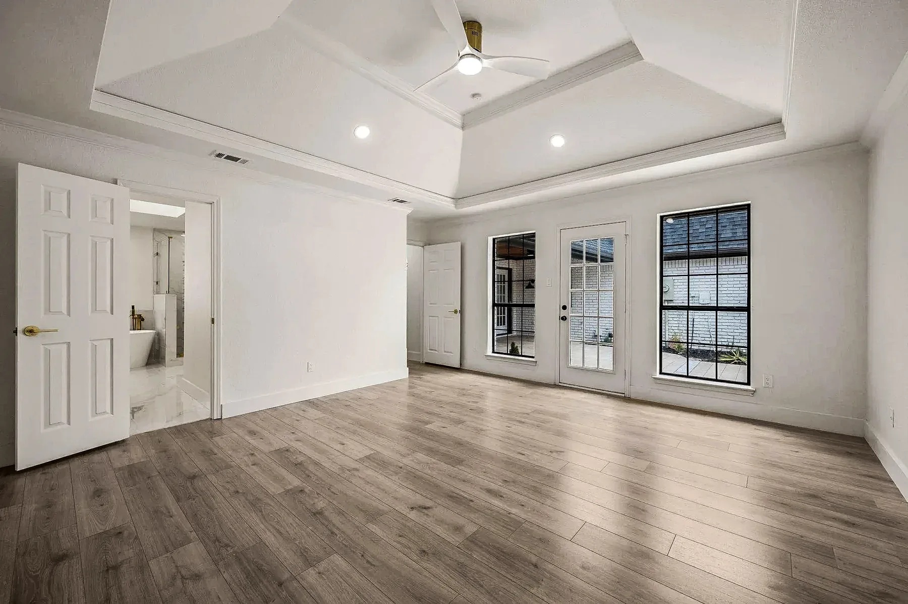 Grapevine primary suite with tray ceiling, black-framed windows, wood-look flooring, and bathroom entry.