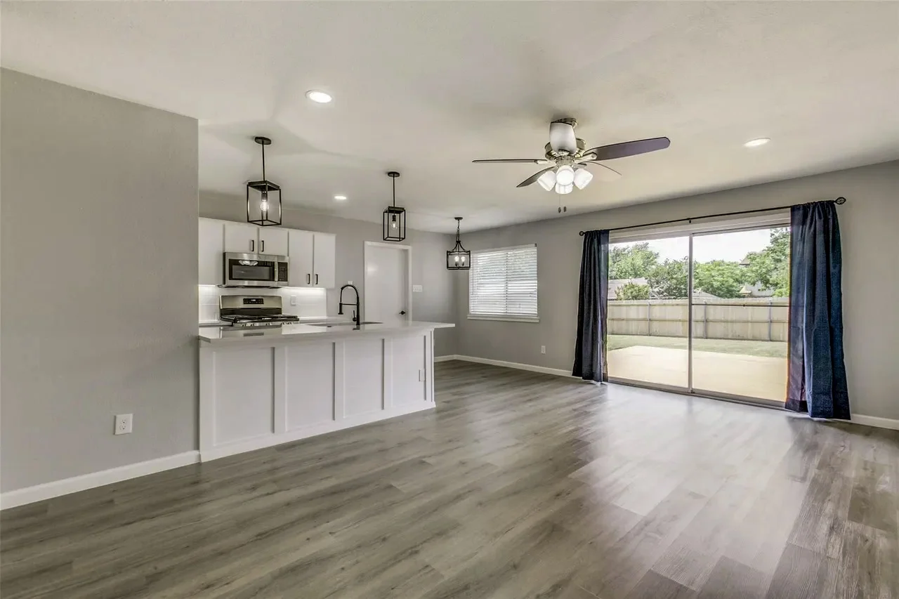 Open living and kitchen area with gray wood-look flooring, white cabinetry, pendant lights, and sliding doors to the backyard in Garland, Texas.