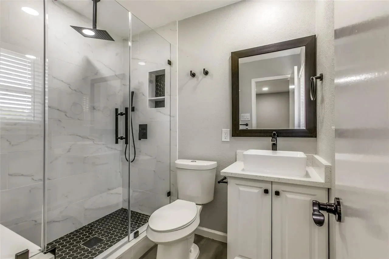 Primary bathroom shower with marble-look tile, frameless glass, black fixtures, pebble-look floor, and white vanity in Garland, Texas.