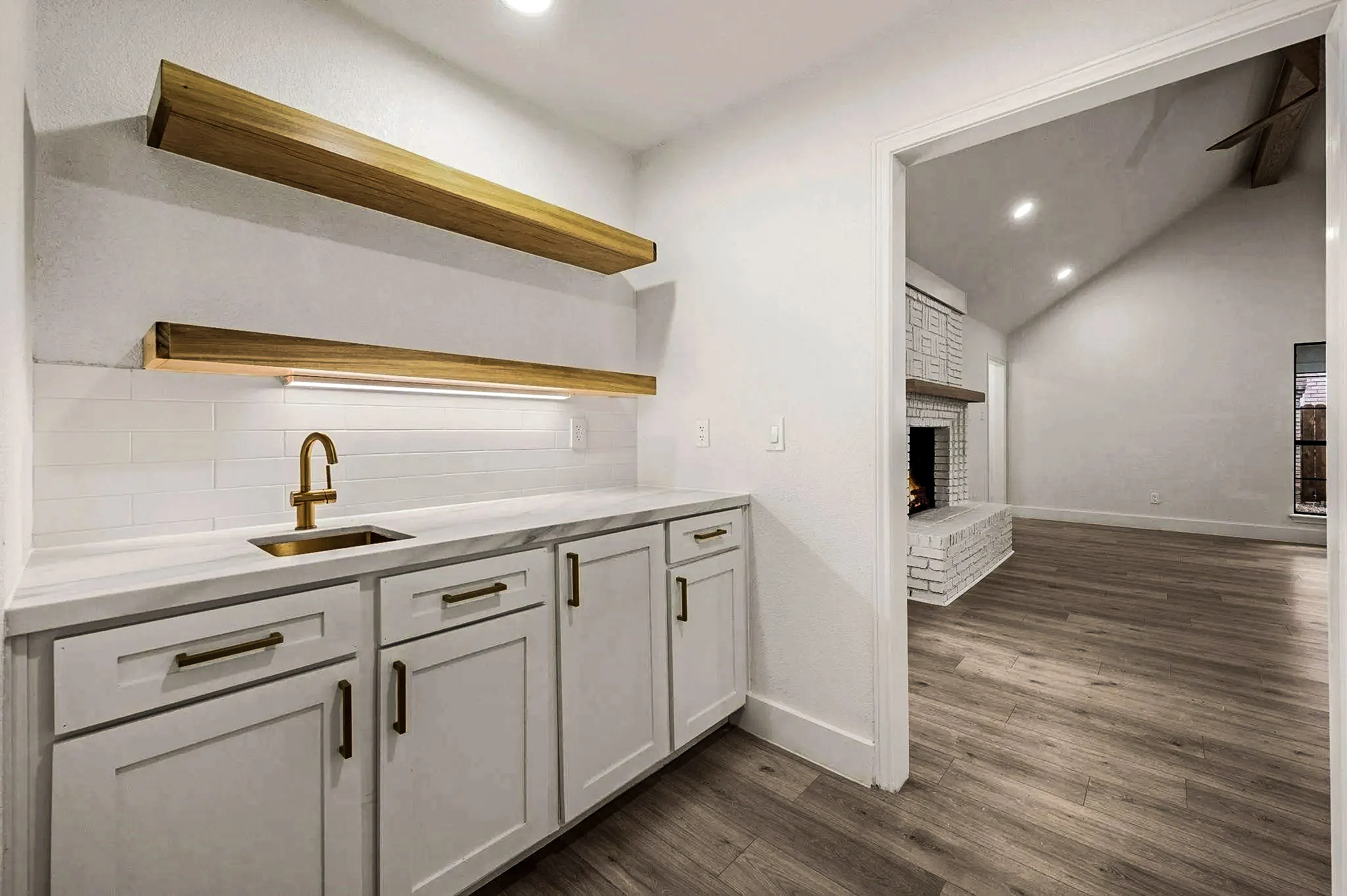 Custom wet bar with white cabinetry, brass faucet, floating brass shelves, stone counters, and wood-look flooring in Grapevine, Texas.