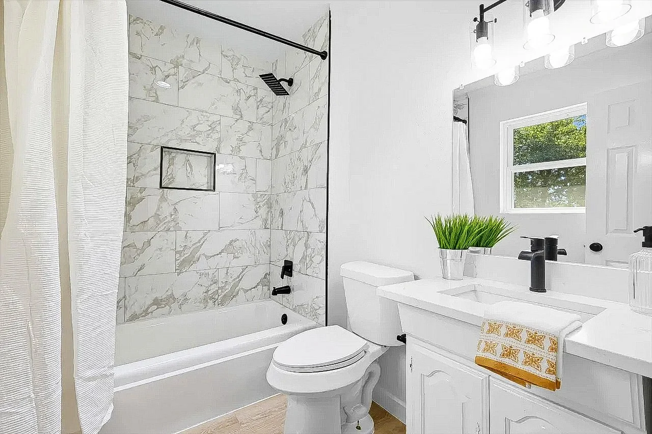 Primary bathroom with marble-look tub surround, white vanity, black fixtures, bright lighting, and neutral flooring in Lewisville, Texas.
