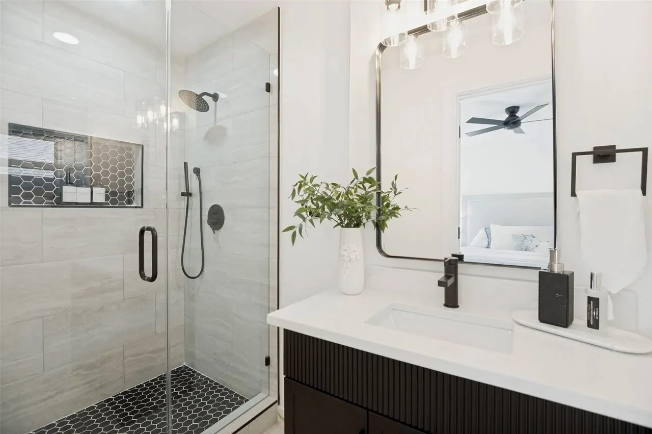 Primary bathroom with a glass shower, gray tile, black fixtures, dark fluted vanity, and illuminated mirrors in Lower Greenville, Dallas, Texas.
