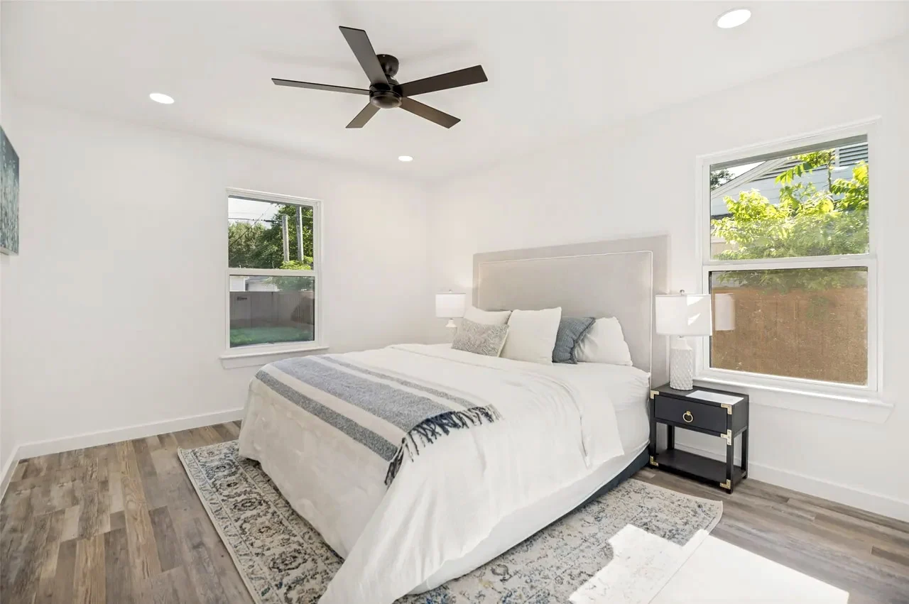 Secondary bedroom with white walls, wood-look flooring, black ceiling fan, corner windows, and staged furnishings in Lower Greenville, Dallas, Texas.