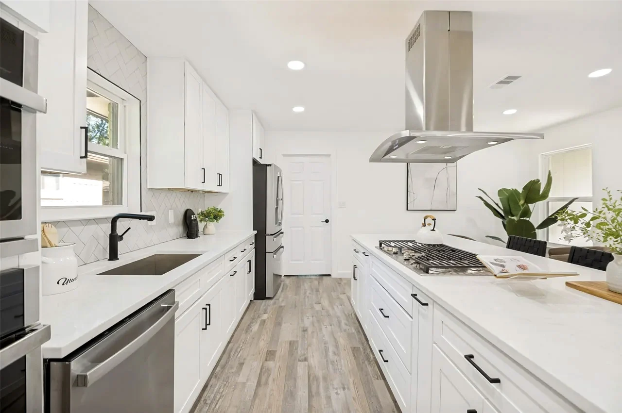 White shaker kitchen with a stainless vent hood, gas cooktop, black faucet, stone counters, and light wood-look flooring in Lower Greenville, Dallas, Texas.