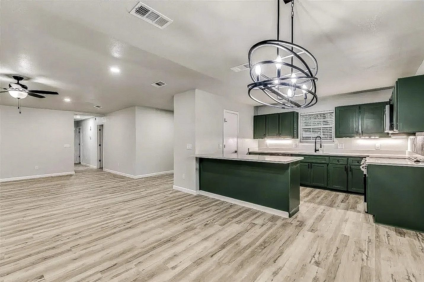 Open kitchen and living area with dark green cabinetry, white counters, geometric pendant, and wood-look flooring in Plano.