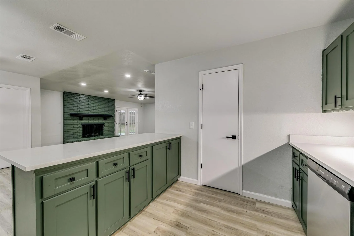 Green kitchen island and cabinetry opening to the living room with a matching green fireplace and light wood-look flooring in Plano, Texas.