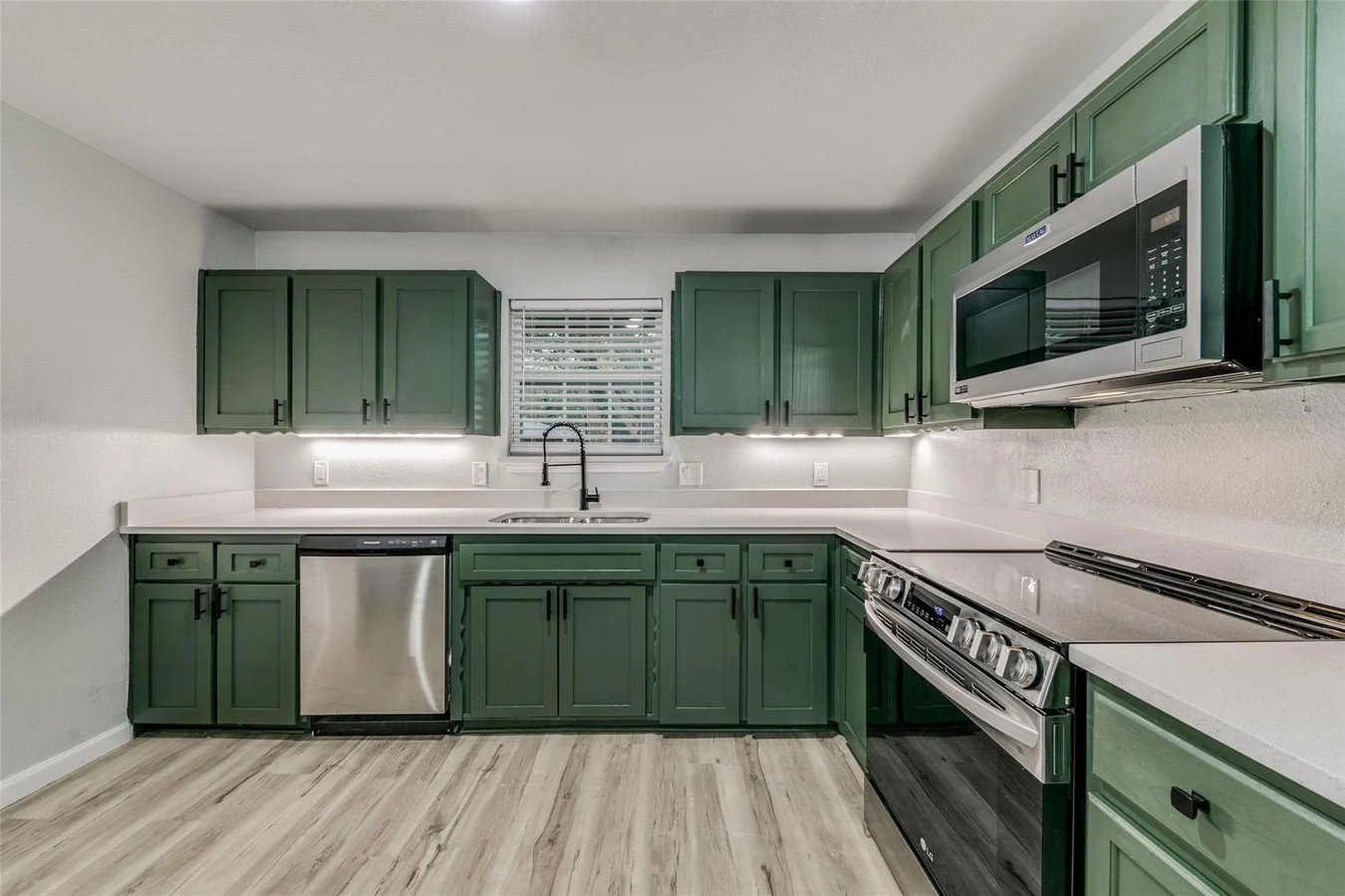 Deep green kitchen with stainless steel range, white counters, black faucet, under-cabinet lighting, and white backsplash in Plano, Texas.