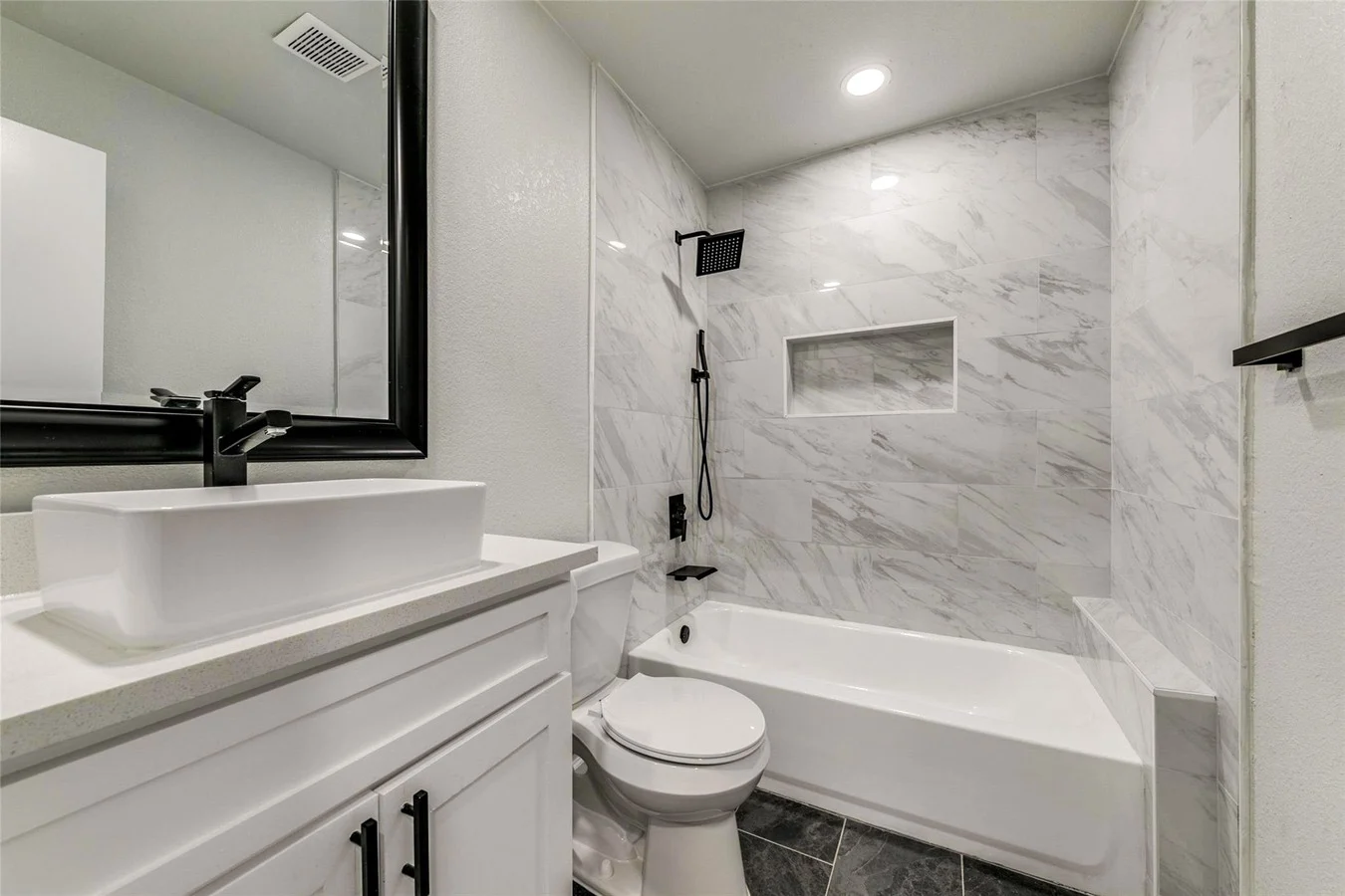 Guest bathroom with a marble-look tub surround, white vessel sink, black fixtures, gray vanity, and recessed lighting in Plano, Texas.