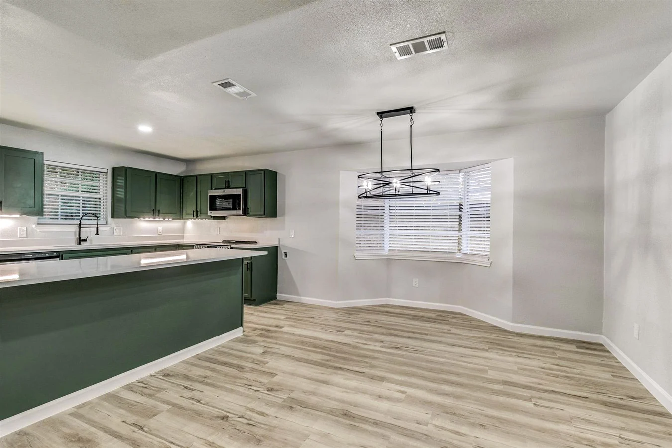 Open dining and kitchen area after wall removal with a green island, modern chandelier, white counters, and light flooring in Plano, Texas.