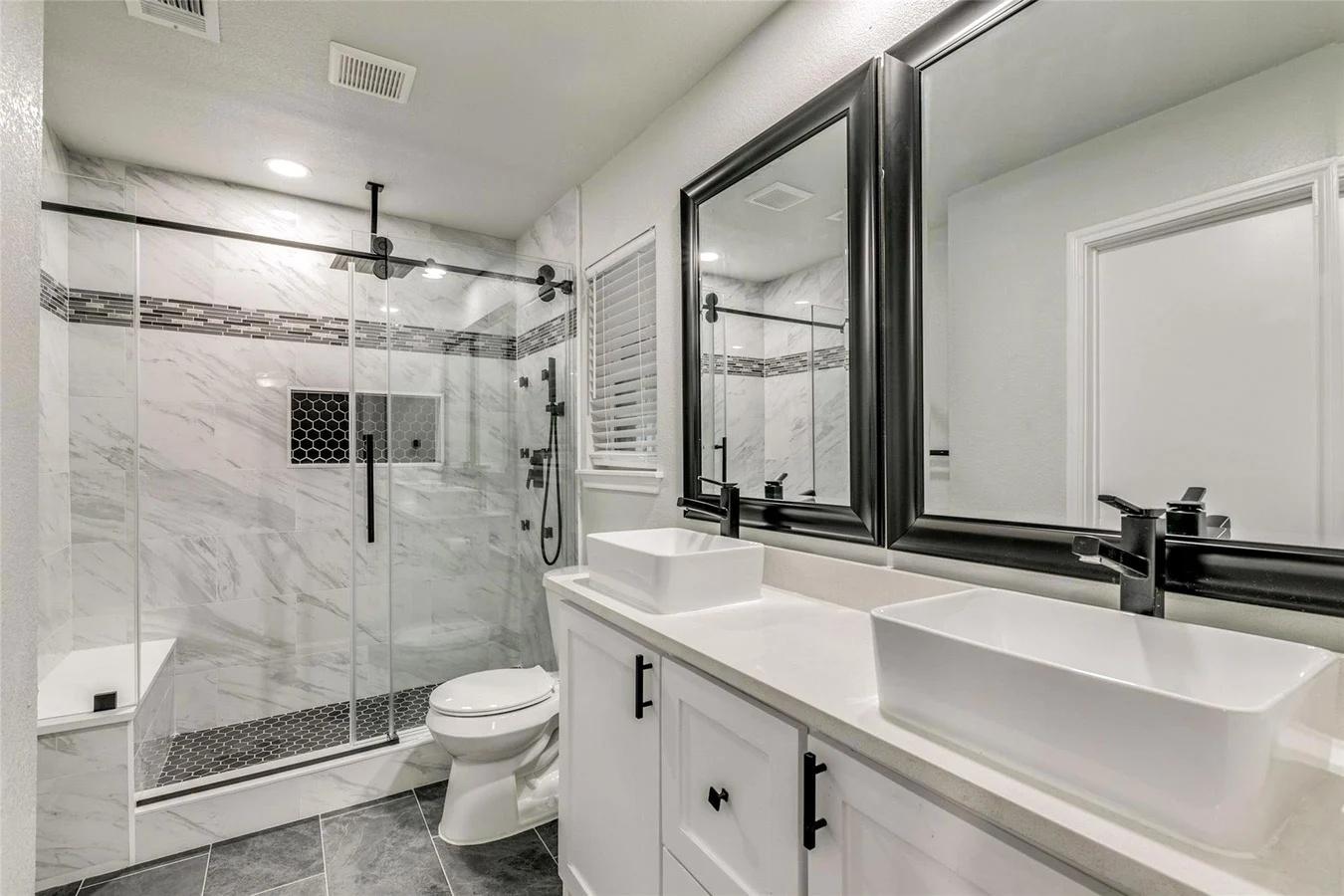 Primary bathroom with a frameless glass shower, marble-look tile, double vessel vanity, black fixtures, and broad mirrors in Plano, Texas.