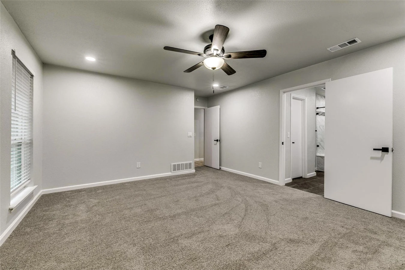 Primary bedroom with new gray carpet, light gray walls, ceiling fan, white trim, and adjacent bathroom access in Plano, Texas.