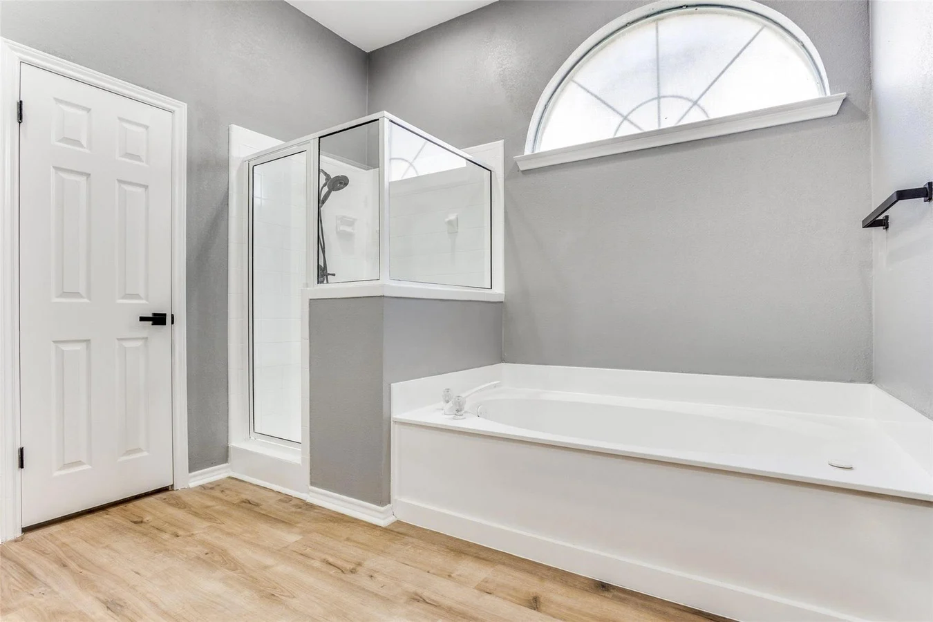 Primary bathroom with a white soaking tub, glass shower, gray walls, arched window, and light wood flooring in Rowlett, Texas.