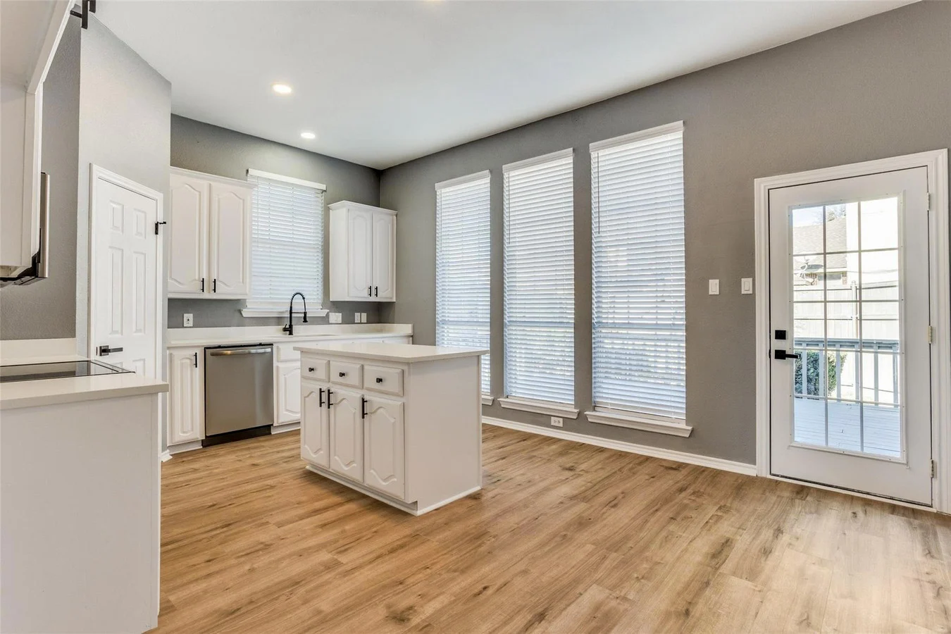 White kitchen with a compact island, stainless appliances, light wood flooring, tall windows, and doors to the rear deck in Rowlett, Texas.